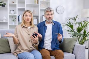 Portrait of disappointed cheated family, man and woman mature couple sitting sad on sofa and looking at camera, using bank credit card and phone for online shopping on the internet. photo