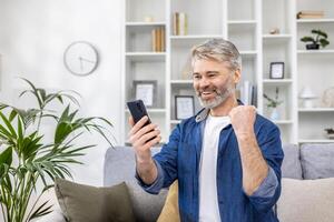 Mature gray haired man alone at home using phone and celebrating victory success happy holding hand up, adult person sitting on sofa in living room on sunny day. photo