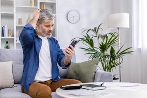 Senior mature adult man sitting on sofa alone at home, using smartphone in living room, reading bad news online from app on phone, upset and nervous. photo