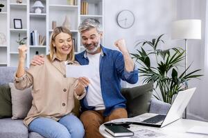 Happy family couple, mature adult man and woman sitting at home on sofa, celebrating victory holding hands up satisfied with family budget, behind paper work family with laptop. photo