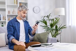 Mature adult man upset and confused reading bad news online from phone, gray haired person sitting on sofa in living room at home working with documents home finances and budget on paper work. photo