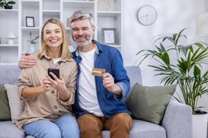 retrato de maduro casado Pareja marido y esposa mujer sentado en sofá en vivo habitación y mirando a cámara, satisfecho en línea compradores de en línea almacenar, participación banco crédito tarjeta y teléfono. foto