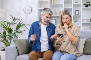 trastornado y engañado pareja, hombre y mujer decepcionado sentado juntos en sofá en vivo habitación, utilizando en línea compras aplicación en teléfono, participación banco crédito tarjeta, negarse dinero transferir. foto