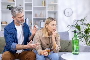 Upset couple sitting in living room on sofa, alcoholic woman drinking a lot of strong alcohol alone, man trying to stop her, discuss everything, relationship problems, alcohol addiction. photo