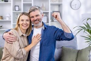 A couple of affectionate senior people in love are sitting on the sofa in their own living room and holding the key to a new home, moving to a new apartment, buying their own house. photo