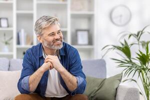 retrato de un de miedo hermoso canoso hombre en un azul camisa sentado a hogar en el sofá y sonriendo mirando a el lado, cruce su brazos en frente de a él. foto