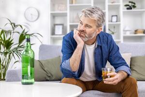 Close up of adult mature man lonely depressed drinking strong alcohol sitting on sofa in living room on day. photo