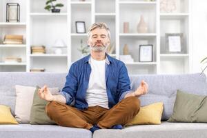 Adult mature man alone at home relaxing sitting in lotus position on sofa, gray haired person relaxing meditating indoors in living room in casual clothes. photo