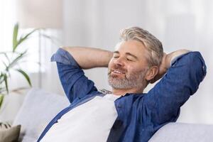 Close-up photo. Senior gray-haired man sitting relaxed on sofa at home and resting, napping. Throwing his hands behind his head, he smiles. photo