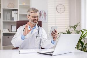Cheerful doctor communicates and consults clients remotely, man in white medical coat works inside medical office, sits at table with laptop, uses tablet computer for call. photo
