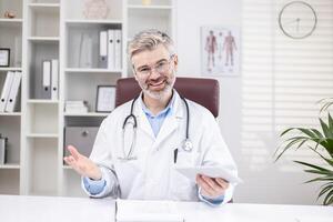 Portrait of a smiling senior male doctor who remotely consults patients online, sits in the office at the table and talks to the camera on a call photo