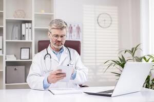 Serious thinking doctor working inside medical office, senior doctor in white medical coat using tablet computer, sitting at table. photo