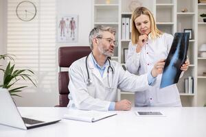 Serious mature doctors thinking and discussing patient's x-ray, man and woman in medical white coats working inside clinic office, working consult. photo