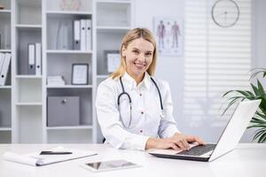 A young female doctor sits in the office in a white coat and with a stethoscope and works on a laptop. Smiling looking at the camera. photo