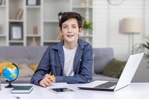Portrait of happy teenage boy student sitting at table at home, studying via distance learning, doing homework, smiling, looking at camera. photo