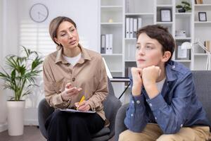 Pensive teenage guy at an appointment with a female psychologist, sitting in a modern office, immersed in his thoughts, consulting a psychotherapist, teenage problems bullying at school. photo