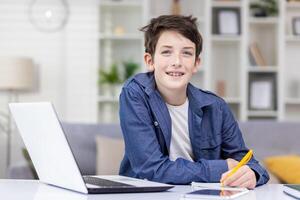 Smiling student boy sitting at a table in a bright room, studying remotely, doing homework, writing in a notebook, smiling, looking at the camera. photo