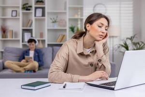 Tired businesswoman working on laptop at home remotely, holding head with hand, tapping on keyboard, programming, mother at work with child in background. photo