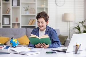 alegre chico colegial lee un libro en su habitación a el mesa, lo hace tarea, trabajos con un tutor en línea en un computadora portátil, distancia aprendiendo. foto