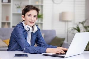 Smiling teenage guy with headphones around his neck, sitting at laptop in bright room, tapping on keyboard, studying, watching , chatting online, looking at camera. photo