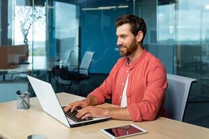 Distance Learning. Portrait of a young male student in a red shirt. Sitting at the desk in the office, studying online on a laptop. Writes a diploma, a course, an abstract, a lecture. photo