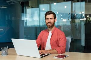 Portrait of a smiling and successful mature man in the office, a businessman with a beard and a red shirt is smiling and looking at the camera, a programmer is using a laptop at work, a creative photo