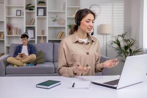 Businesswoman in headset sitting at table in living room in front of laptop, having call, attractive woman working in call center, working from home, mother working with child in background. photo