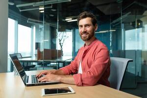 Portrait of a smiling and successful mature man in the office, a businessman with a beard and a red shirt is smiling and looking at the camera, a programmer is using a laptop at work, a creative photo