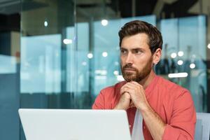 Portrait of a young thoughtful serious man businessman, freelancer, programmer sitting at a table, working on a laptop, folded his hands together, looks to the side. photo