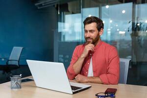 Portrait of a young male programmer, designer, freelancer in a red shirt who sits and works in the office at a laptop. He looks thoughtfully at the monitor, talks on a call. photo