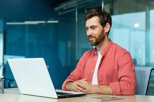 Mature adult businessman working on laptop inside office, programmer smiling contentedly reading and checking software code, man in red shirt. photo