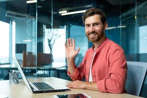 Portrait of successful financier businessman inside office at workplace, man smiling and looking at camera waving hand gesture of greeting, small business owner working using laptop photo