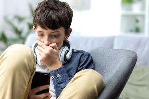 Young teenager boy reading bad news on phone, son sitting on sofa in living room with headphones at home closeup. photo