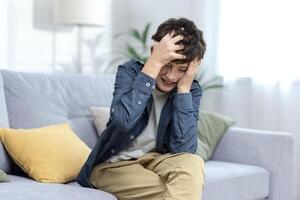 A child, a boy, a teenager, sits alone at home on the sofa and holds his head in his hands. Suffers from headache, tired, panic attack, personality traits, domestic violence. photo