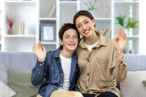 Portrait of a family, a mother and a teenage son sitting at home on the sofa and hugging, waving at the camera, greeting each other, chatting on a call. photo