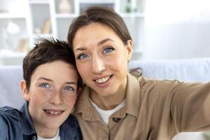 Son and mother at home sitting on the sofa in the living room, looking at the camera, smiling, taking a selfie photo, and talking via call online. photo