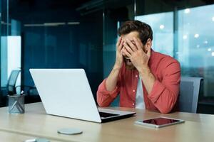 Problems at work. A young man sits in the office at a table with a laptop. Upset and tired, he holds his face with his hands. Screams with anger. photo