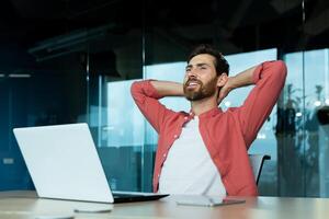 Businessman successfully finished work, man with hands behind head rests at workplace, programmer smiles and looks at window while sitting at desk photo