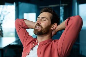 Close-up photo. Young man freelancer, student sits on a chair in the office, coffee shop and rests. He put his hands behind his head, closed his eyes. relaxes, meditates, dreams. photo