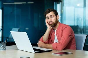 Boring office work. A young man sits in the office at a table with a laptop, leans on his arm and is bored, tired, looks to the side. Professional burnout, depression. photo