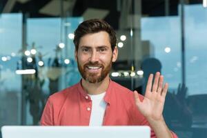 Portrait of a young male programmer in a red shirt working in the office at a laptop. He looks at the camera with a smile, waves his hand. Close-up photo. photo