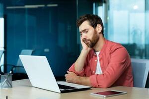 Overtime, deadline. Tired young male programmer, freelancer sits in the office and tries to work at the laptop. Bored, he sits at the table and rests his head on his hand. photo