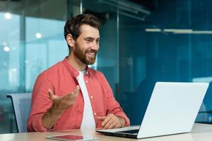 Learning online. A young male teacher in a red shirt sits in the office at the table, works on a laptop, teaches through a call, conference, webinar. Explains, smiles. photo