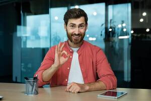 A young businessman conducts an online business training, webinar. Sitting in the office, talking on a call, recording a podcast, pointing to the camera with his hand ok. photo