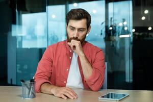 Shocked and upset young man sitting at the desk in the office, thoughtfully holding his head with his hand. Fired, downsizing, salary reduction, unemployed, company closing. photo