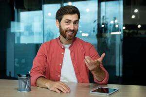 Portrait of a smiling young man sitting at the desk in the office in front of the camera and talking via call, conducting business training, online conference, webinar, business meeting. photo