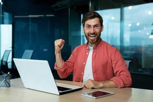 Happy young student sitting in office, campus in front of .aptop, showing victory success gesture with hand, celebrating, looking at camera, smiling. photo