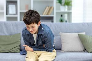 A child, a boy, a teenager, sits alone at home on the sofa and holds his stomach. Suffering from pain, bent over, needs medical help. photo