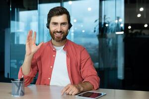 Portrait of a young man in a red shirt and headset. He sits at the desk in the office, looks at the camera, waves his hand, smiles. photo