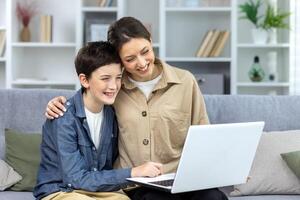 Mother and son hugging together sitting on sofa at home in living room watching online using laptop, family spending time together watching online educational course. photo